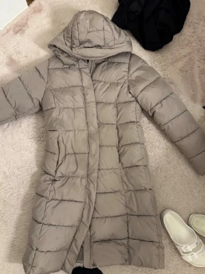 Lång beige dunjacka med huva - Snygg lång beige dunjacka med huva och quiltad design. Jackan har raka ärmar, dold dragkedja och två sidofickor. Perfekt för kalla dagar när du vill hålla dig varm och stilren.