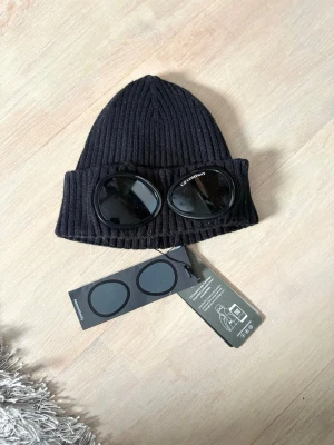 Svart C.P. Company goggle-mössa - Säljer en svart ribbstickad mössa från C.P. Company med de ikoniska goggle-linserna framtill. Mössan är tillverkad i mjukt material och har en tight passform. Perfekt för dig som vill ha en streetwear-inspirerad look med unik detalj.