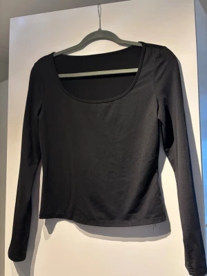 Svart långärmad topp med rund hals - En stilren svart långärmad topp med bred, rundad urringning. Toppen är figurnära och har en enkel, minimalistisk design som passar till det mesta. Tillverkad i ett stretchigt och mjukt material för skön passform. Skims material på tröjan 
