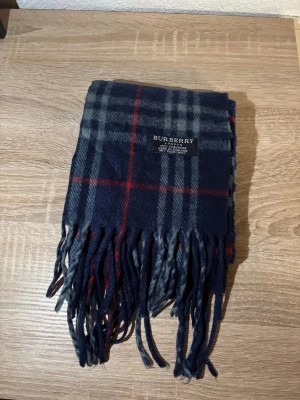 Rutig marinblå halsduk från Burberry - Snygg halsduk från Burberry i marinblått med klassiskt rutmönster i grått och rött. Gjord i mjuk kashmir, med fransar i ändarna och diskret Burberry-logga. Perfekt för att ge din outfit en cool och lyxig touch.