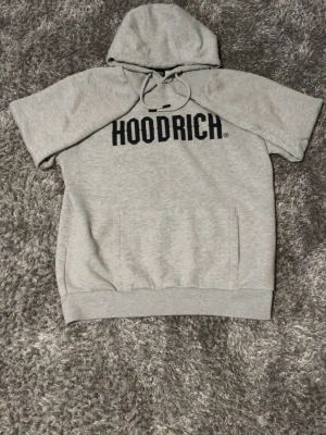 Grå HOODRICH hoodie med kort ärm - Snygg ljusgrå hoodie från HOODRICH med korta ärmar och stor svart logga på bröstet. Hoodien har huva med dragsko och en stor magficka framtill. Tillverkad i mjukt sweatshirtmaterial, perfekt för en avslappnad streetwear-look.