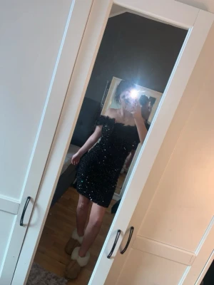 Svart glittrig offshoulder klänning - Snygg svart kortklänning med glittriga detaljer och offshoulder-modell. Klänningen har en figurnära passform och volangdetaljer vid axlarna som ger en extra touch. Perfekt för dig som vill sticka ut och glänsa. Är i storlek L men skulle mer säga M. Bara använd en gång!