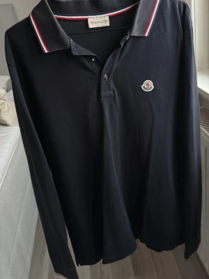 Svart långärmad piké från Moncler - Snygg svart långärmad pikétröja från Moncler med klassisk krage och knappar framtill. Köpt på NK.  Den har två små larm hål men inget som syns