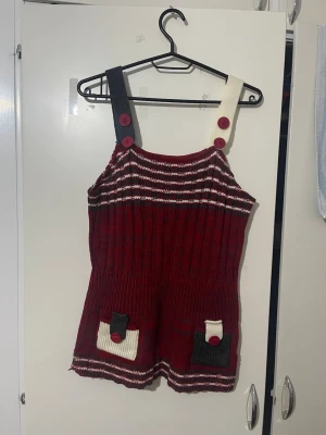 Röd stickad topp med fickor och halsduk  - Unik stickad topp i vinrött med vita och mörkgröna detaljer. Den har två olika färgade axelband med stora knappar och två små fickor framtill. Randiga detaljer upptill och nedtill ger en cool retrovibe. Perfekt för dig som vill sticka ut! En halsduk följer med också, den är lite sliten men går fortfarande att använda. (det går att köpa toppen utan eller med halsduken).