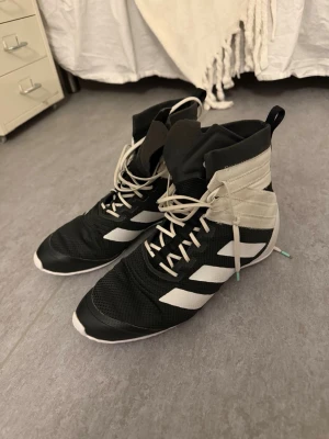 Adidas boxningsskor svart/vit - Snygga boxningsskor från Adidas i svart och vitt med klassiska tre ränder på sidan. Skorna har högt skaft, snörning hela vägen upp och är tillverkade i mesh och syntetmaterial för bra ventilation och lätt känsla. Perfekta för träning eller match.
