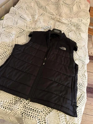 North Face Väst - Svart väst från The North Face med dragkedja framtill och två sidofickor. Västen har en lite skråma på vänster axel