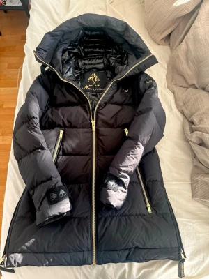 Svart dunjacka från Moose Knuckles - Snygg svart dunjacka från Moose Knuckles med guldfärgade dragkedjor och detaljer. Jackan har stor huva, två snedställda fickor med dragkedja och logga på ärmen. Fodrad med dun för extra värme och har en längre modell som skyddar mot kyla.