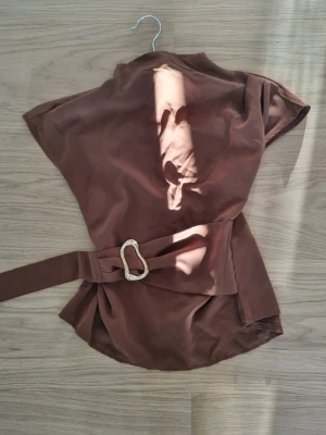 Brun omlottopp med bälte - Snygg brun omlottopp med tillhörande bälte och dekorativt spänne i midjan. Toppen har korta ärmar och en avslappnad passform som ger en cool och trendig look. Perfekt att styla med jeans eller kjol för en chic outfit.