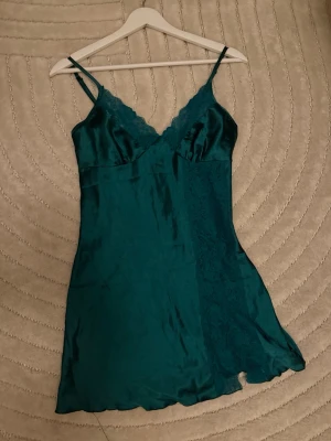 Mörkgrönt satinnattlinne med spets - Snyggt mörkgrönt nattlinne i satin med tunna axelband och spetsdetaljer vid bysten även en liten slitt i spetsen längst ner. Nattlinnet har en lätt utsvängd passform och en mjuk, glansig yta som känns lyxig mot huden. Perfekt för mysiga kvällar hemma.