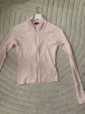 Ljusrosa ziptröja  - Snygg ljusrosa ziptröja med hög krage och långa ärmar. Tröjan har figurnära passform och är tillverkad i ett stretchigt, lätt material som passar perfekt till träning eller som ett extra lager. Dragkedja framtill. 