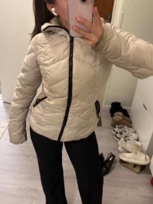 Beige dunjacka med huva - Säljer en beige dunjacka med huva och svarta dragkedjor. Jackan har två snedställda fickor med dragkedja och quiltat mönster. Perfekt för kalla dagar och riktigt skön att bära.