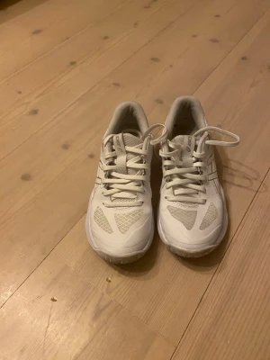 Vita sneakers från Asics - Säljer ett par fräscha vita sneakers från Asics med klassisk design. Skorna har snörning, meshpaneler för bra ventilation och detaljer i syntetmaterial. Perfekta för dig som gillar sportig stil och vill ha bekväma skor till vardags.