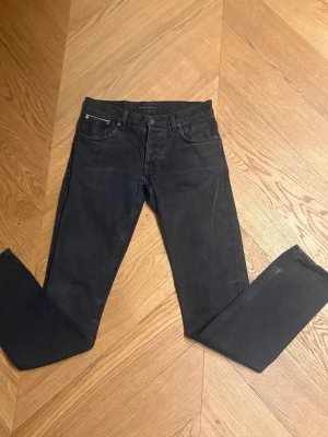 Svarta slim fit jeans från Nudie  - Snygga svarta slim fit jeans från Nudie med klassisk femficksmodell och knappgylf. Jeansen har raka ben och diskreta sömmar, perfekta för en clean och stilren look. Materialet är mjukt och stretchigt för extra komfort.