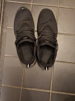 Svarta Nike sneakers i textil - Säljer ett par helsvarta Nike sneakers med klassisk låg profil och rund tå. Skorna har en ovandel i mesh-liknande textil, svarta snören och diskret Nike-logga på sidan. Sulan är svart och det finns en liten ögla bak för enkel påtagning.  SKA TVÄTTAS ORDENTLIGT OCH PAKETET FINNS TROLIGTVIS, SKRIV DM