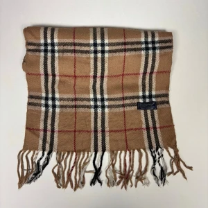 Beige rutig vintage halsduk från Burberrys - Klassisk beige vintage halsduk från Burberrys med ikoniskt rutmönster i svart, vitt och rött. Halsduken har fransar i ändarna och är tillverkad i mjukt material som känns lyxigt och stilrent. Perfekt accessoar för att lyfta din outfit.