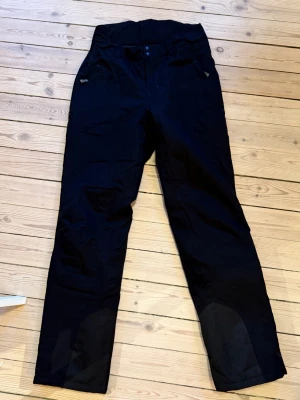 Svarta täckbyxor från Neomondo - Svarta täckbyxor från Neomondo med raka ben och förstärkta partier vid knän och benslut. Byxorna har dragkedjor vid fickorna och justerbara benslut. Sparsamt använda för skidåkning