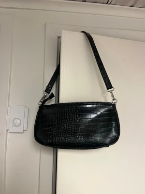 Svart axelväska från Even&Odd - Snygg svart axelväska från Even&Odd i croco-mönstrat konstläder. Väskan har en avlång form, silverfärgade detaljer och dragkedja upptill. Perfekt för dig som gillar trendiga accessoarer med lite edge.