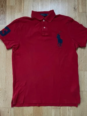 Röd pikétröja från Polo Ralph Lauren - Klassisk röd pikétröja från Polo Ralph Lauren med blå logga broderad på bröstet och en stor blå siffra 3 på ärmen. Tröjan har krage, knappar vid halsen och är tillverkad i mjuk bomull. Perfekt till en avslappnad men ändå stilren look.