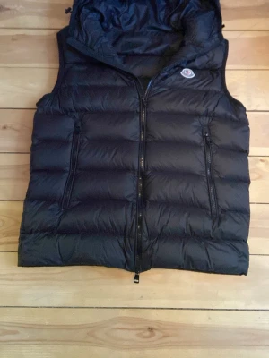 Svart dunväst från Moncler - Snygg svart dunväst från Moncler med klassisk logga på bröstet. Västen har två dragkedjeförsedda fickor framtill och en hög krage. Tillverkad i glansigt/puffer material och fylld med dun för extra värme. Perfekt för lager-på-lager och streetstyle. Jag köpte västen av en kompis så jag vet inte om den är äkta men jag tror 99% att den är det eftersom kartonen ser bra ut och den har scan, loggan ser bra ut och size taggen står i siffror och även väldigt bra kvalitet! 