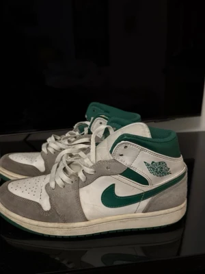 Air Jordan 1 Mid vit/grön/grå - Snygga Air Jordan 1 Mid sneakers i vitt, grönt och grått. Ovandelen är i en mix av läder och mocka med gröna detaljer på swoosh och krage. Klassisk Jordan Wings-logga på sidan och perforerad tåbox för extra stil. Perfekta för dig som gillar streetwear och retrovibbar.