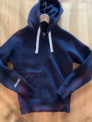 Mörkblå zip hoodie från Russemerch - Mörkblå hoodie från Russemerch med dragkedja framtill, vita snören i huvan och kängurufickor. Klassisk modell med ribbade muddar och diskret Russell-brodyr på ärmen. Perfekt för chill dagar och enkel att matcha med jeans eller joggers.