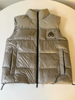 Beige dunväst från Moose Knuckles - Helt nytt skick nypriset är 8000tusen säljer den just nu för 3749 priset är inte fast i sten!!                          Snygg beige dunväst från Moose Knuckles med hög krage och svart dragkedja. Västen har två framfickor och en bröstficka med logotypdetalj. Tillverkad i glansigt material och perfekt för lager-på-lager under kalla dagar.