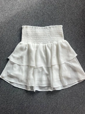 Vit volangkjol från Chelsea - Supersöt vit kortkjol från Made by Chelsea med smock i midjan och dubbla volanglager. Tillverkad i ett lätt material som ger en snygg siluett. Använd bara ett fåtal gånger.