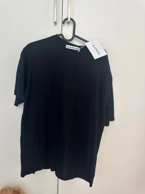 Svart t-shirt från Acne Studios - En klassisk svart t-shirt från Acne Studios med rund hals och avslappnad passform. Tillverkad i mjuk bomull och har en enkel, stilren design utan mönster eller tryck. Perfekt basplagg för alla tillfällen.