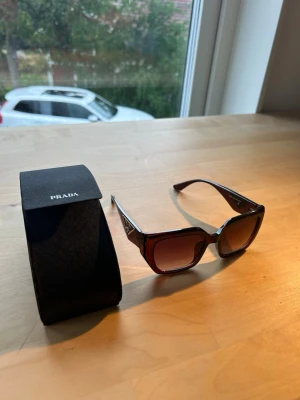 Bruna fyrkantiga solglasögon från Prada - Snygga solglasögon från Prada med fyrkantig form och mörkbruna bågar. Glasögonen har en tydlig logga på sidorna och kommer med originalfodral. Perfekta för att lyfta din stil med en lyxig touch. 100% uv skydd
