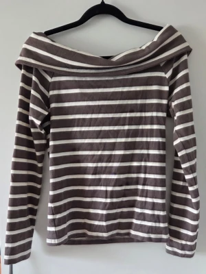 Randig offshoulder topp brun/vit - Säljer en brun och vit randig offshoulder topp med långa ärmar. Toppen har en bred krage som vilar över axlarna. Perfekt för dig som gillar en avslappnad men ändå trendig stil.