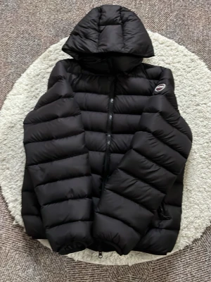 Colmar svart dunjacka med huva - Snygg svart dunjacka med huva och quiltad design. Jackan har dragkedja framtill och en diskret logga på bröstet. Perfekt för kalla vinterdagar och riktigt skön att bära. Flera praktiska fickor och elastiska muddar vid ärmsluten.