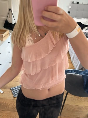 Ljusrosa oneshoulder topp med volanger - Säljer en superfin ljusrosa oneshoulder topp med flera lager volanger. Toppen är ärmlös och har en romantisk vibe, perfekt för dig som gillar söta detaljer. Materialet känns lätt och luftigt, vilket gör den skön att bära under varma dagar.