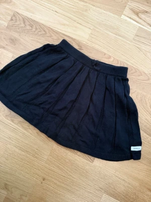 Svart stickad kjol från Newbie - Mysig svart stickad kjol från Newbie med bred resår i midjan och diskreta vertikala ränder. Kjolen har en klassisk kort modell och är perfekt för en avslappnad och trendig look.