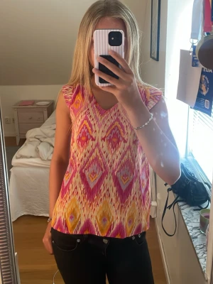 Mönstrad ärmlös topp från ONLY - Färgglad ärmlös topp från ONLY med v-ringning och ett iögonfallande mönster i rosa, orange och gul. Perfekt för varma dagar och ger en riktigt somrig vibe. Lätt och luftig modell som sitter snyggt och bekvämt.