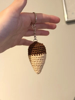 Handvirkad glassnyckelring - Söt handgjord nyckelring formad som en glass i strut, virkad i brunt och beige garn. Perfekt att hänga på väskan eller nyckelknippan för en lekfull touch. Metallkedja och ring för enkel fastsättning.