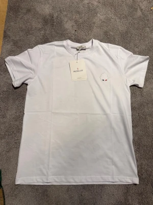 Vit t-shirt från Moncler - Snygg vit t-shirt från Moncler med diskret broderad logga på bröstet. Klassisk passform och rund halsringning. Tillverkad i mjuk bomull som känns skön mot huden. Perfekt för en clean och stilren look.