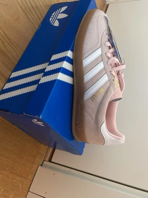 Adidas Gazelle rosa sneakers - Snygga Adidas Gazelle sneakers i ljusrosa mocka med vita detaljer och klassiska tre ränder på sidan. Gummisula i brunt och guldfärgad text på sidan. Skön och stilren modell som passar till det mesta. Perfekt för dig som gillar retrovibbar och streetstyle. Nya och helt oanvända 
