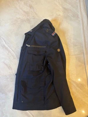 Fieldjacket från Snoot - Snygg mörkblå fieldjacket från Snoot med flera praktiska fickor och dragkedja framtill. Jackan har en klassisk design med diskreta detaljer och en liten logotyp på ärmen. Perfekt för blåsiga dagar och enkel att matcha med olika outfits. 