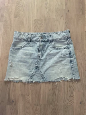 Ljus jeanskjol med rå kant - Trendig ljusblå jeanskjol med rå, fransig kant och slitna detaljer. Kjolen har klassiska fem fickor, bälteshällor och knappgylf. Snygg söm framtill och broderade bakfickor ger extra stilpoäng. Perfekt för en avslappnad look.