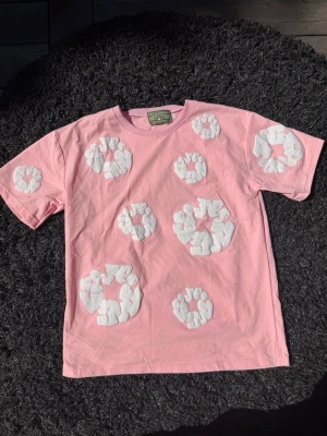 Rosa t-shirt från Denim Tears - Säljer en rosa t-shirt från Denim Tears med vita blommiga applikationer på framsidan och ärmarna. T-shirten har en klassisk passform och är gjord i mjuk bomull. Perfekt för dig som vill sticka ut med en unik och trendig look.