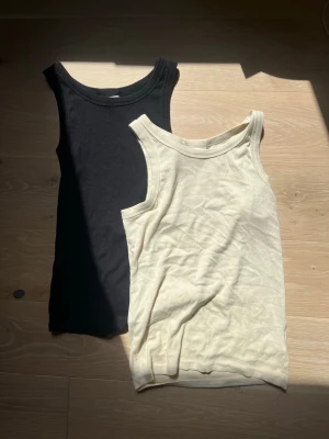 2-pack ribbade linnetoppar  - Två ribbade linnetoppar  i svart och beige. Båda är ärmlösa med rund halsringning och tillverkade i mjuk bomull. Knappt andvända och säljer pågrund av att dem aldrig kommer till andvändning. 