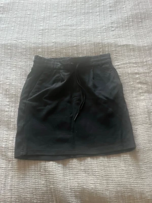 Svart kortkjol med snörning - En svart kortkjol med snörning i midjan och enkel design. Kjolen har raka linjer och är perfekt för en avslappnad och stilren look. Passar till många olika outfits och är lätt att matcha.
