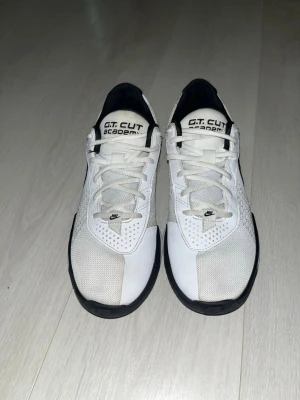 Nike G.T. Cut Academy vita sneakers - Säljer ett par vita Nike G.T. Cut Academy sneakers med svarta detaljer och meshpaneler för extra ventilation. Skorna har klassisk snörning, låg profil och en bekväm sula som passar perfekt till streetwear eller sportiga outfits. Bara använda några fåtal gånger inomhus. Ny pris är runt 1000kr. Priset går att diskutera.