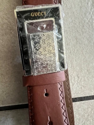 Brunt skärp med silverdetaljer från Gucci längd 114 cm - Snyggt brunt skärp från Gucci i skinn med präglad logga på remmen och ett stort silverspänne med GG-mönster. Perfekt accessoar för att lyfta din outfit med en exklusiv touch.