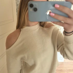 Beige off shoulder stickad tröja - Trendig beige tunn stickad tröja med off shoulder-design och hög hals. Mjuk och skön, perfekt för dig som vill ha en stilren men ändå unik look. Långa ärmar och avslappnad passform gör den enkel att matcha till olika outfits.