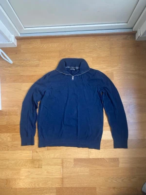 Mörkblå stickad tröja Jack & Jones - Mörkblå stickad tröja från Jack & Jones med halv dragkedja och hög krage. Tröjan har lång ärm och ribbade muddar vid ärmslut och nederkant. Perfekt för lager-på-lager och enkel att matcha med jeans eller chinos.