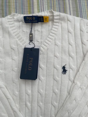 Vit kabelstickad tröja Polo Ralph Lauren - Kabelstickad vit tröja från Polo Ralph Lauren med V-ringning och klassisk marinblå logga broderad på bröstet. Tröjan har långa ärmar och ribbade muddar vid ärmslut och nederkant. Perfekt för en clean och stilren look.