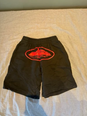 CORTEIZ shorts - Säljer ett par svarta shorts med elastisk midja och en stor röd tryckdetalj framtill. Shortsen har en bakficka med knapp och är tillverkade i mjukt bomullsmaterial. Perfekta för sommaren och chill dagar.