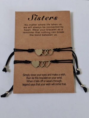 Systerskap armband med hjärtan - 2 stycken matchande systerskap- och vänskapsarmband fästa på ett tillhörande gåvokort med en fin text. Dessa kallas ofta för "Wish Bracelets" (önskearmband) och är tillverkade av flätat svart nylonrep med justerbara knutar samt en silverfärgad metallberlock i form av sammanlänkade hjärtan.