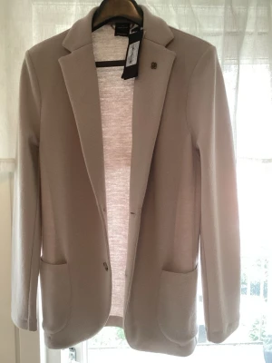 Beige kavaj från Gran Sasso - Stilren beige kavaj från Gran Sasso i mjuk travel wool. Klassisk modell med två framfickor, enkel knappstängning och snyggt slag. Perfekt för dig som vill ha en clean och modern look. Liten metallogo på bröstet ger extra detalj. Kavajen är aldrig använd och prislappen sitter kvar (5995kr).